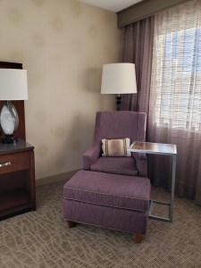 Purple chair and ottoman by window, in hotel room, striped throw pillow on seat, side table on the right, dresser with lamp on the left.