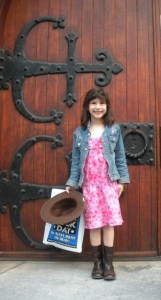 Girl standing in front of a wooden door with wrought-iron accents. She is wearaing a pink flowered dress, a denim jacket, unbuttoned, and cowboy boots. She is holding in her right hand a cowboy hat and a comic book bag. She is smiling the forced smile of a preschooler.