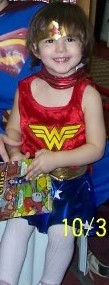 Toddler dressed as Wonder Woman, holding and reading a comic book, smiling.